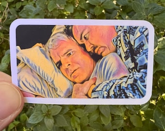 May include: A rectangular sticker featuring a stylized illustration of two individuals in a close embrace. One person with light hair rests their head on a pillow, while the other person leans in closely. The artwork uses warm tones and bold outlines.
