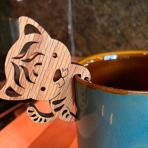 May include: A wooden cat-shaped tea bag holder on a blue ceramic mug. The mug has a brown interior and a Yosemite National Park emblem. The tea bag holder is made of light brown wood with cut-out details. The mug is a light blue colour.