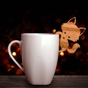 May include: A white ceramic mug with a wooden fox cutout attached. The fox is light brown and appears to be climbing on the mug. The background is dark with blurred golden lights, set on a dark wooden surface.