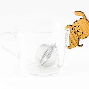 May include: A clear glass teacup with a wooden tea infuser shaped like a dog. The infuser has a chain attached to it and is hanging over the edge of the cup. The infuser is in the shape of a dog with its tongue sticking out.