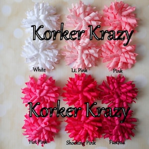 May include: A collection of Korker Krazy hair bows in various shades of pink and white. The bows are arranged in a cluster, with the colors labeled as White, Lt. Pink, Pink, Hot Pink, Shocking Pink, and Fuschia. The text "Korker Krazy" is displayed in a black font.