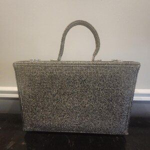 May include: A rectangular handbag featuring a textured black and white tweed-like fabric exterior. The bag has a single arched top handle with a matching woven pattern and silver-toned metal hardware attachments at the top corners of the frame.