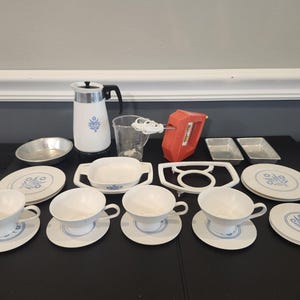 May include: A collection of kitchenware including a white coffee pot with blue floral patterns, a clear measuring cup, an orange electric hand mixer, two small metal loaf pans, a round metal pan, and several white ceramic cups and saucers.