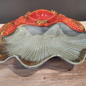 May include: A ceramic serving dish with a crab design. The dish is light blue with a textured, fan-like pattern. The crab is red with gold accents on the claws and body. The dish is on a wooden surface.