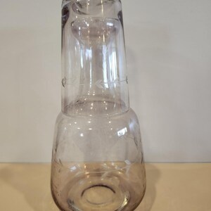 May include: A clear glass water carafe with a matching glass. The carafe has a bulbous base and a narrow neck, with a decorative etched leaf pattern. The glass is inverted on top of the carafe.