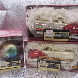 May include: Three Christmas ornaments in boxes. Two boxes contain silver and red train ornaments labeled "1950 Lionel Santa Fe". The third box holds a round ornament with a grape design. All ornaments are Hallmark Crown Reflections.