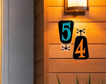 May include: Black and teal metal house numbers 5 and 4 mounted on a wooden wall. The number 5 is teal with a black background, and the number 4 is black with an orange background. A vintage-style light fixture is above the numbers.