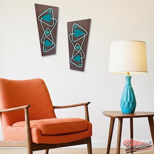 May include: Two mid-century modern wall art panels. Each panel is a dark brown, elongated shape with a teal geometric design. The panels are mounted on a white wall. An orange chair and a teal lamp are also in the image.