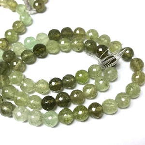 May include: A strand of faceted green gemstone beads. The beads are round and have a slightly irregular shape. The beads are strung on a white thread.