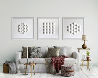May include: Three framed white geometric wall art pieces above a white sofa with patterned pillows and a red and black throw blanket. A small side table with a lamp and a decorative stool complete the living room setting.