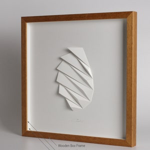 May include: A white paper sculpture of geometric shapes framed in a natural wood box frame. The frame has a white mat and white side profiles.