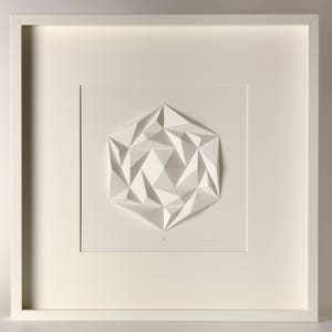 May include: A white framed print of a 3D geometric design. The design is made of paper and is folded into a complex pattern. The print is signed by the artist.