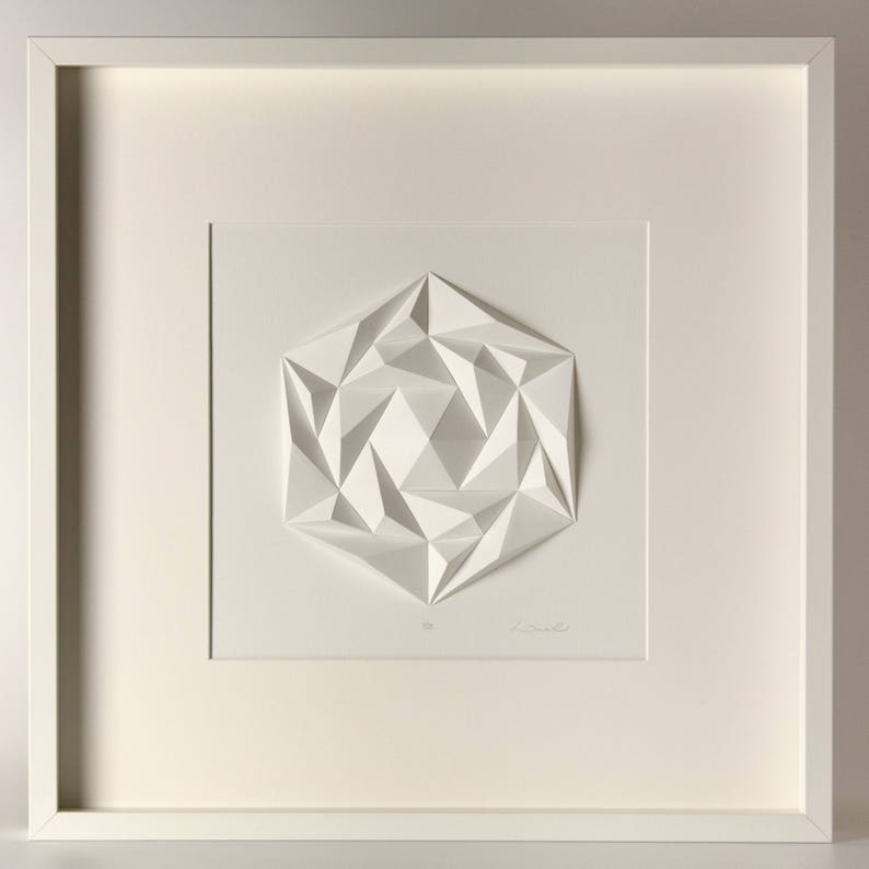 May include: A white framed print of a 3D geometric design. The design is made of paper and is folded into a complex pattern. The print is signed by the artist.
