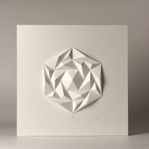 May include: A white paper sculpture of a geometric shape, resembling a flower or star, with a textured, three-dimensional design. The sculpture is mounted on a white background.