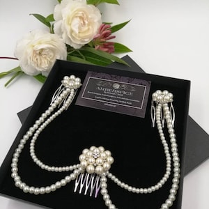 May include: A hair accessory set featuring three silver-toned metal combs adorned with clusters of white faux pearls and small clear crystals. The combs are connected by two draped strands of white faux pearls, presented in a black box with a business card.