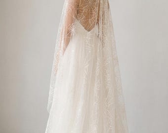 May include: A floor-length white wedding veil featuring an intricate, embroidered spiderweb pattern throughout the sheer fabric. The veil is attached at the crown and drapes over the shoulders and back, extending to the floor with a circular train.