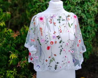May include: A sheer, white capelet with delicate floral embroidery in shades of pink, red, and green. The capelet features a scalloped lace trim along the edges and is displayed on a white mannequin.