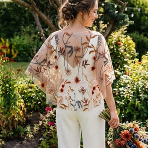 Puede incluir: Un mono blanco con una capa superior transparente de manga corta, decorada con bordados de flores silvestres y follaje en tonos óxido, marrón y azul. La capa tiene una espalda en forma de V y un ajuste fluido sobre el mono blanco sin mangas.