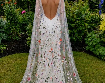 May include: A white wedding dress with a low back and a sheer, floral-embroidered cape. The dress is adorned with colorful flowers, and the cape drapes to the ground. The image is taken outdoors in a garden setting.