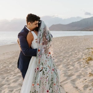 Può includere: Una coppia si abbraccia sulla spiaggia. La sposa indossa un abito da sposa bianco con ricami floreali e un velo abbinato. Lo sposo è in abito blu navy. Lo sfondo presenta una spiaggia sabbiosa, l'oceano e montagne.