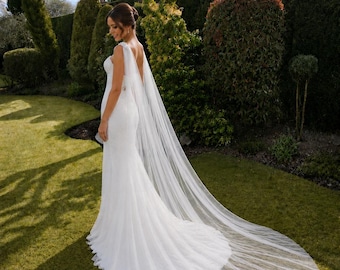 May include: A white wedding dress with a long train. The dress features a fitted bodice and a tulle skirt. The bride is in a garden with green grass and trees. The veil extends behind the dress, creating a dramatic effect.