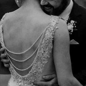 May include: A sleeveless gown featuring a deep scoop back adorned with three draped strands of white pearls. The bodice and shoulder straps are embellished with intricate silver beadwork and sequins, complemented by a row of small fabric buttons.