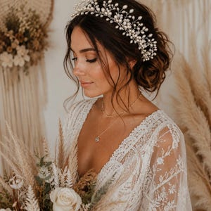 Puede incluir: Una novia con una corona nupcial de perlas y cristales. La corona es plateada y está adornada con pequeñas perlas y cristales. Lleva un vestido de encaje blanco y sostiene un ramo de flores.