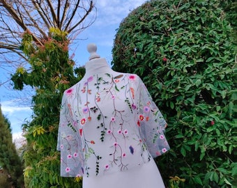 May include: A sheer, cropped top featuring intricate, multi-colored floral embroidery. The design showcases delicate stems, leaves, and various small blossoms in shades of pink, red, orange, and purple scattered across the translucent fabric.