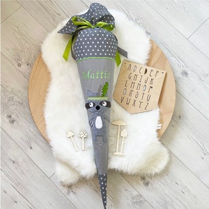 Könnte beinhalten: Ein grauer und weißer Schultüte mit einem Waschbär-Motiv und dem Namen "Mattis" bestickt. Die Tüte ist mit grünem Band und weißen Sternen verziert. Im Hintergrund befindet sich ein Holzalphabet-Brett.