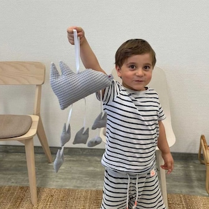 Könnte beinhalten: Ein Kind hält ein graues und weißes Plüschtier mit einer hängenden Schnur aus kleineren grauen und weißen Plüschtieren. Das Kind trägt ein weiß-schwarz gestreiftes Shirt und eine passende gestreifte Hose.