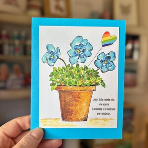 Può includere: Biglietto di auguri con bordo blu con un'illustrazione ad acquerello di fiori blu in un vaso marrone. Un cuore arcobaleno fluttua sopra i fiori. Il testo recita: "Solo un piccolo promemoria che sei qualcosa da celebrare ogni giorno."