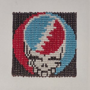 Może przedstawiać: Wiszący obraz z koralików z kolorowym logo Grateful Dead. Logo jest wykonane z czerwonych, białych, niebieskich i czarnych koralików.