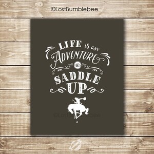 May include: A brown background with white text that reads "Life is an Adventure Saddle Up". A white silhouette of a cowboy riding a horse is below the text.