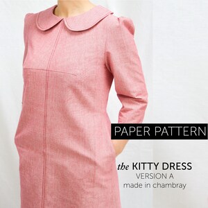 a side view of A woman wearing a red winter dress with 3/4 length sleeves with gathering, round collar, and her hand is in her pocket. The dress has interesting bust dart details and topstitching.