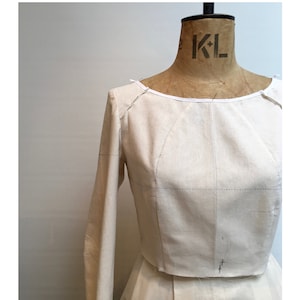 Könnte beinhalten: Ein cremefarbenes Kleidungsstück, möglicherweise ein Oberteil oder ein Kleid, ist an einer Vintage-Schneiderpuppe befestigt. Das Wort "MAVEN" ist oben in Schwarz gedruckt, darunter "the TOILE GUIDE". Die Schneiderpuppe hat "KL" auf dem Hals aufgedruckt.