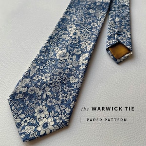 Puede incluir: Una corbata azul con un estampado floral blanco. La corbata se llama "the WARWICK TIE PAPER PATTERN".
