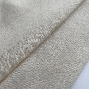 May include: Close-up of a folded piece of natural-colored linen fabric. The fabric has a textured weave, with visible fibers and a slightly uneven surface. The color is a soft, off-white or cream.
