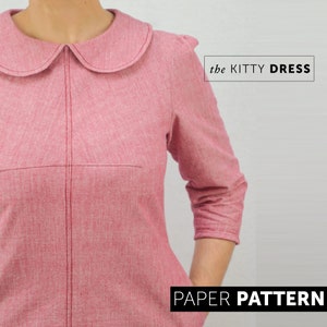 front view of A woman wearing a red winter dress with 3/4 length sleeves, Peter pan collar, and her hand is in her pocket. The dress has interesting bust dart details and topstitching.