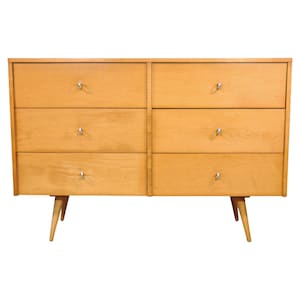 Op de afbeelding: Een mid-century modern dressoir met zes lades. Het dressoir is gemaakt van lichtgekleurd hout en heeft zilverkleurige knoppen. Het heeft taps toelopende poten en een strak, minimalistisch ontwerp.