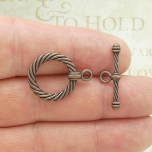 May include: A pair of antique copper-toned metal toggle clasps. The clasp is a twisted rope design with a round ring on one end and a T-shaped bar on the other end.