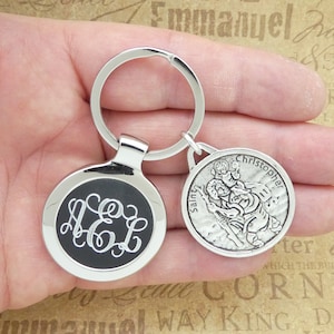 May include: A silver keychain with a black round charm featuring a monogram and a silver charm with a depiction of Saint Christopher. The charm reads "Saint Christopher".