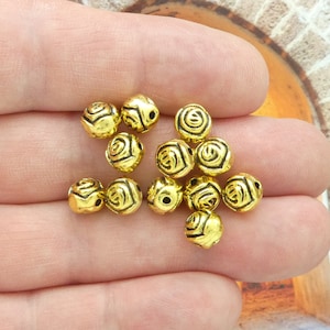 May include: A close-up of a pile of gold-colored metal beads with a rose design. The beads have a hole in the center for stringing.