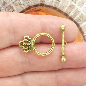 May include: Gold-toned metal toggle clasp with a crown design. The clasp has a round ring and a straight bar with rounded ends.