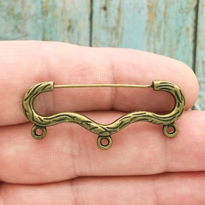 May include: A bronze-colored metal brooch with a wavy, twig-like design. The brooch has a pin and catch closure and three dangling loops.