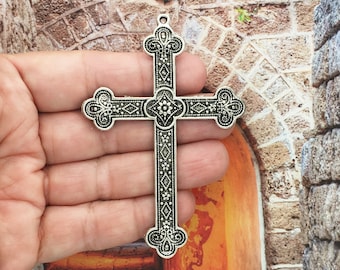 May include: A silver-toned cross pendant with intricate floral designs and a diamond pattern. The cross is ornate and detailed, with a loop at the top for hanging.