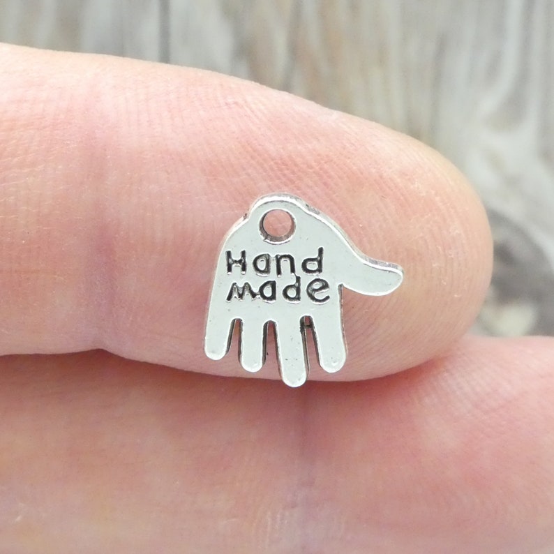 Puede incluir: Encanto de plata en forma de mano con las palabras "Hand Made" inscritas en &eacute;l.