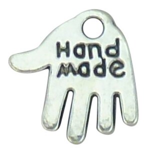 May include: A silver-coloured charm shaped like a hand, with the words "HAND MADE" in capital letters. It features a small hole at the top for attaching to jewellery. The charm is likely made of metal and is suitable for crafting.