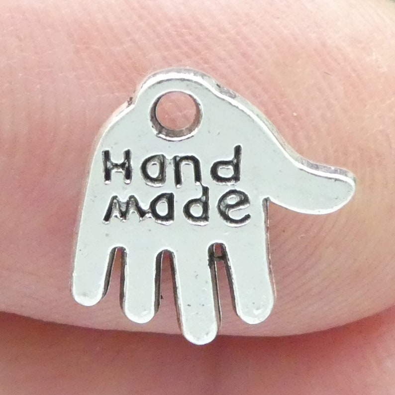 May include: A small, silver-coloured charm in the shape of a hand. The words "Hand Made" are inscribed in bold, black letters across the palm. The charm has a small hole at the top for stringing.