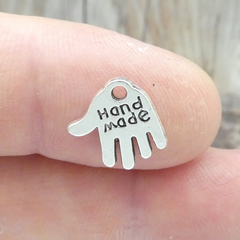 May include: A small, silver-coloured charm in the shape of a hand. The words "Hand Made" are printed in black, block letters on the palm. The charm has a small hole at the top for stringing.