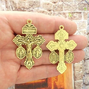 May include: Two gold-toned metal crucifixes with ornate designs. The crucifix on the left features a figure of Jesus Christ on the cross, with three medallions depicting the Virgin Mary. The crucifix on the right has three medallions with religious text, including "Behold This Heart" and "Which Has So Loved Men".